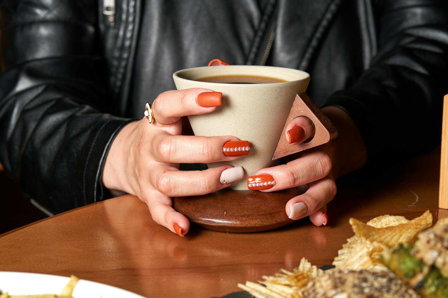 Jesienny manicure 2024: modne kolory, wzory i techniki dobrane do Twojego typu urody Dłonie z jesiennym manicure trzymają kubek kawy przy drewnianym stole