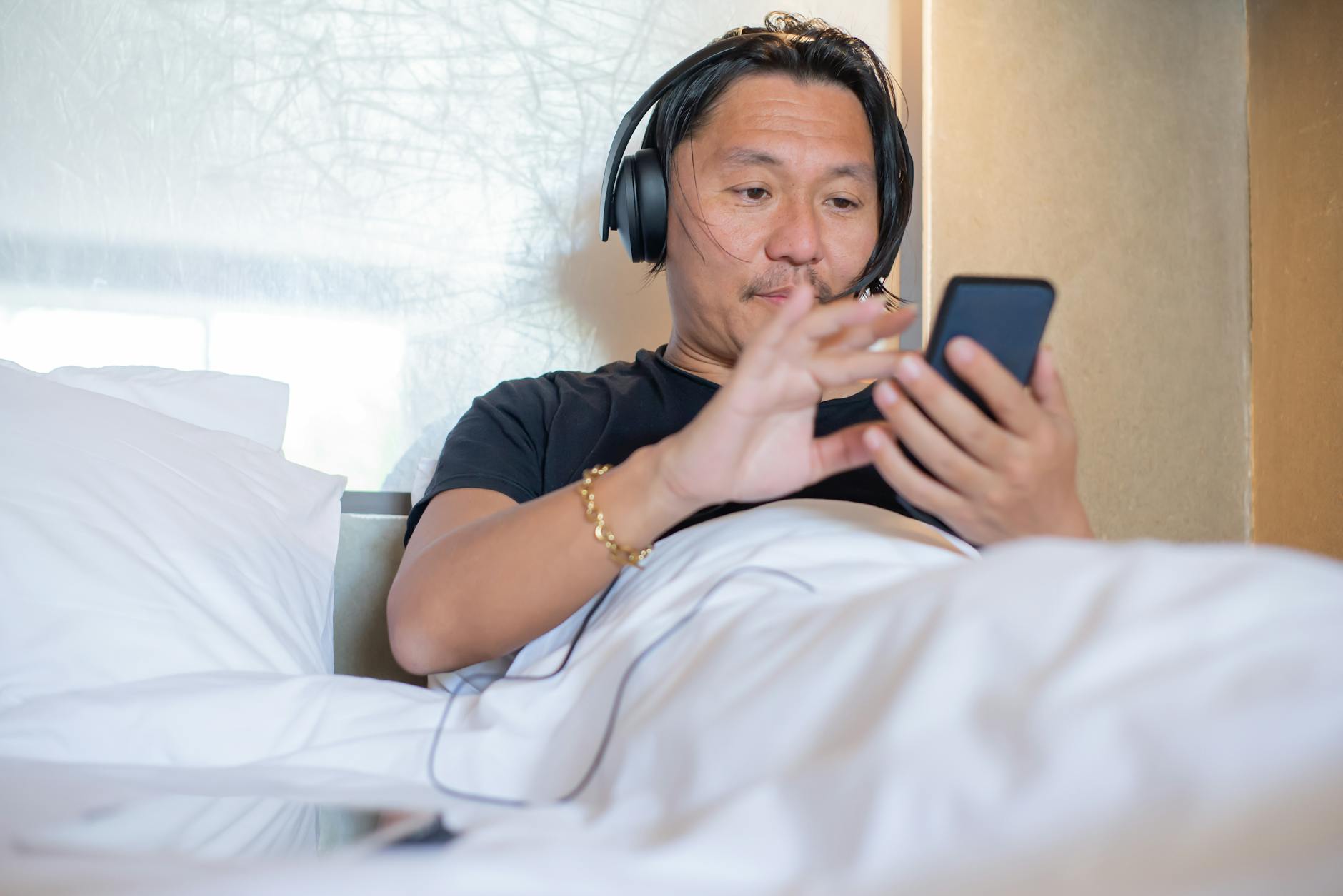 Klasyka literatury w audio: gdzie legalnie znaleźć lektury szkolne z dobrą jakością Mężczyzna azjatyckiego pochodzenia słucha audiobooka na smartfonie w sypialni