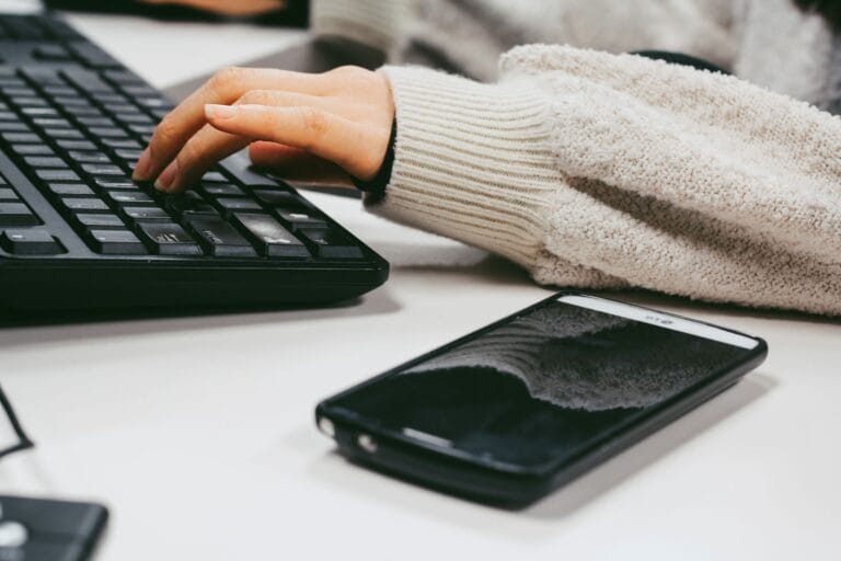 Dłoń kobiety piszącej na klawiaturze obok smartfona na biurku